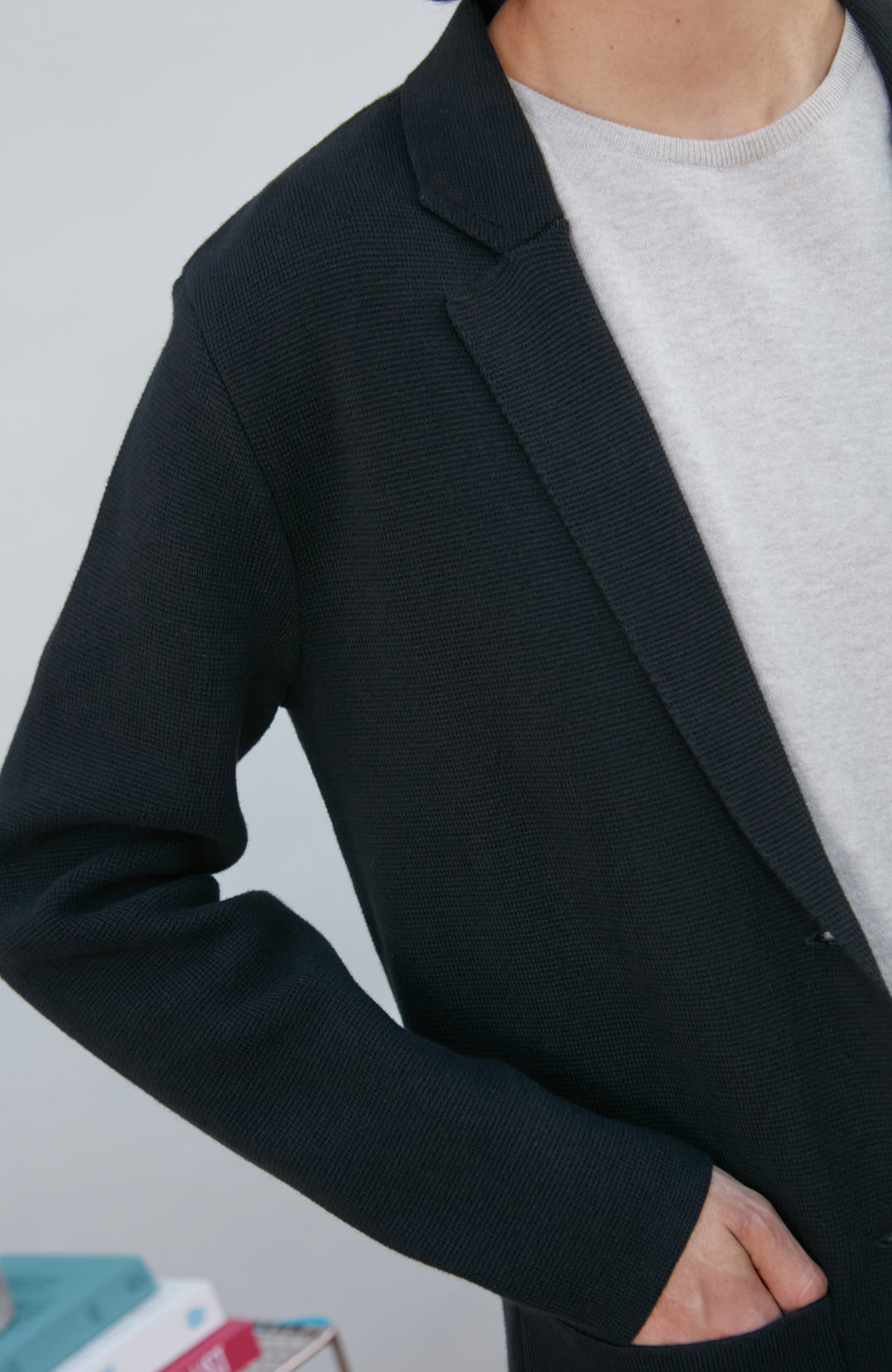 A model wearing a black knitted blazer by Paul James Knitwear over a light gray crewneck t-shirt and olive green pants, standing in a minimalist room with a white wall and a small wire shelf.