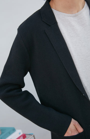 A model wearing a black knitted blazer by Paul James Knitwear over a light gray crewneck t-shirt and olive green pants, standing in a minimalist room with a white wall and a small wire shelf.