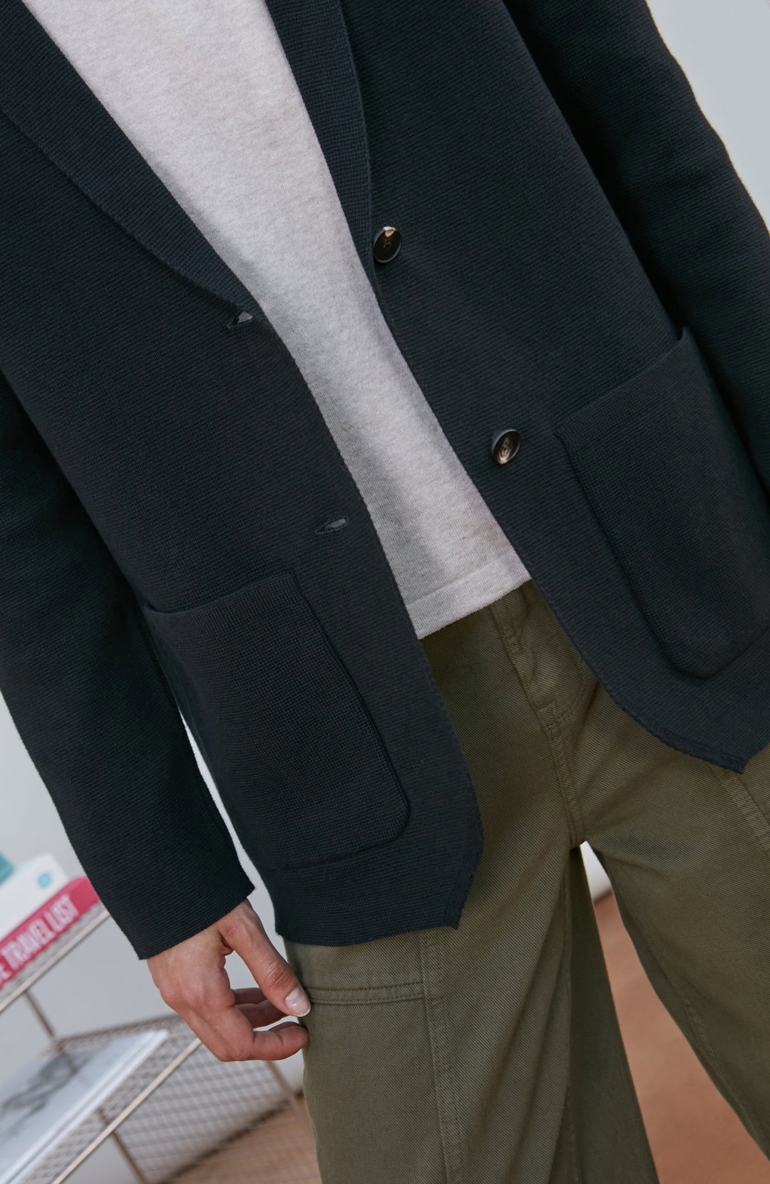A model wearing a black knitted blazer by Paul James Knitwear over a light gray crewneck t-shirt and olive green pants, standing in a minimalist room with a white wall and a small wire shelf.
