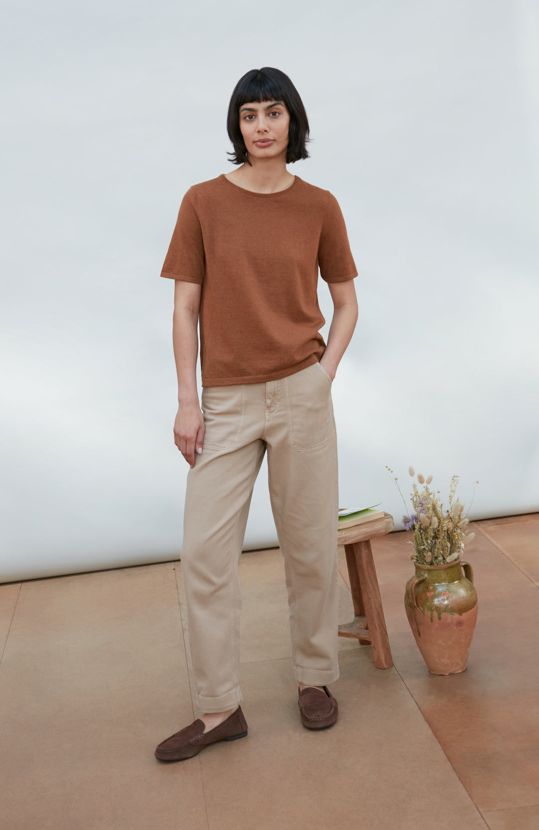 A woman wearing a short-sleeve crewneck knit top in a warm rust brown color By Paul James Knitwear, paired with beige trousers, photographed against a clean off-white studio background.
