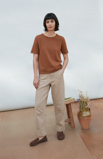 A woman wearing a short-sleeve crewneck knit top in a warm rust brown color By Paul James Knitwear, paired with beige trousers, photographed against a clean off-white studio background.
