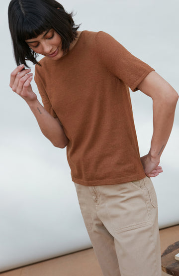 A woman wearing a short-sleeve crewneck knit top in a warm rust brown color By Paul James Knitwear, paired with beige trousers, photographed against a clean off-white studio background.