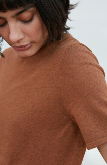 A woman wearing a short-sleeve crewneck knit top in a warm rust brown color By Paul James Knitwear, paired with beige trousers, photographed against a clean off-white studio background.