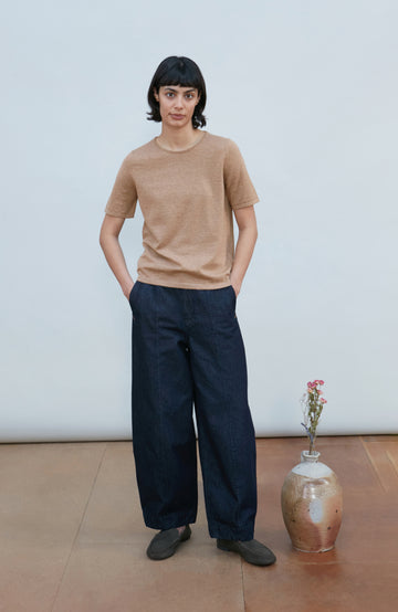A woman with a short dark bob hairstyle wearing a light brown short-sleeve crewneck knit top by Paul James Knitwear and dark navy pants, standing against a plain light gray background.