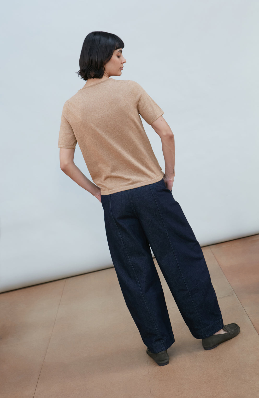 A woman with a short dark bob hairstyle wearing a light brown short-sleeve crewneck knit top by Paul James Knitwear and dark navy pants, standing against a plain light gray background.