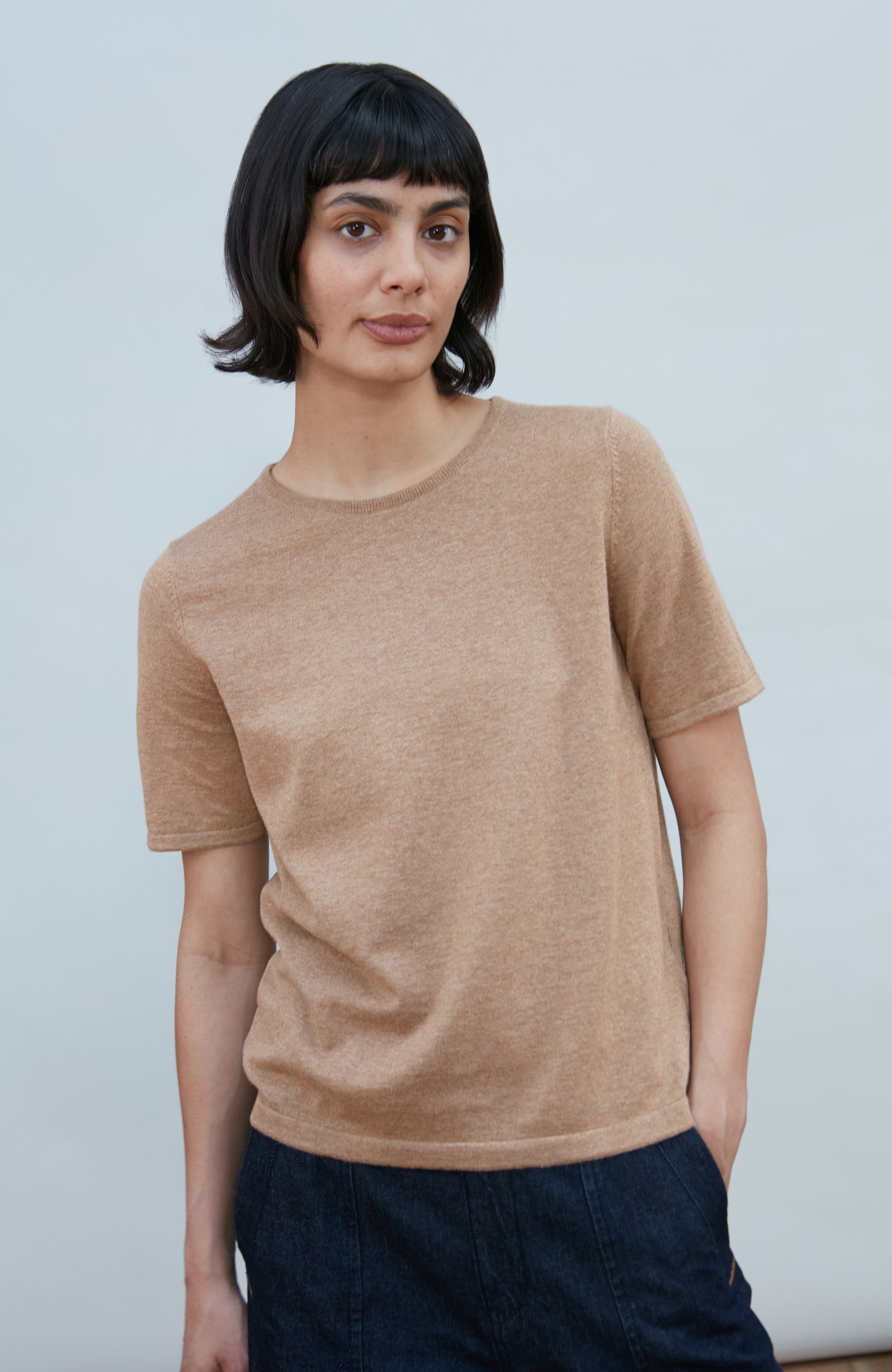 A woman with a short dark bob hairstyle wearing a light brown short-sleeve crewneck knit top by Paul James Knitwear and dark navy pants, standing against a plain light gray background.