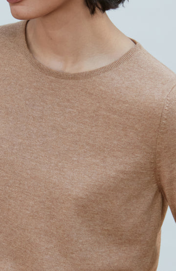A woman with a short dark bob hairstyle wearing a light brown short-sleeve crewneck knit top by Paul James Knitwear and dark navy pants, standing against a plain light gray background.