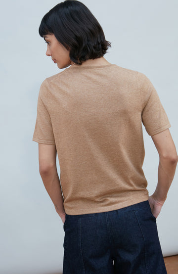 A woman with a short dark bob hairstyle wearing a light brown short-sleeve crewneck knit top by Paul James Knitwear and dark navy pants, standing against a plain light gray background.