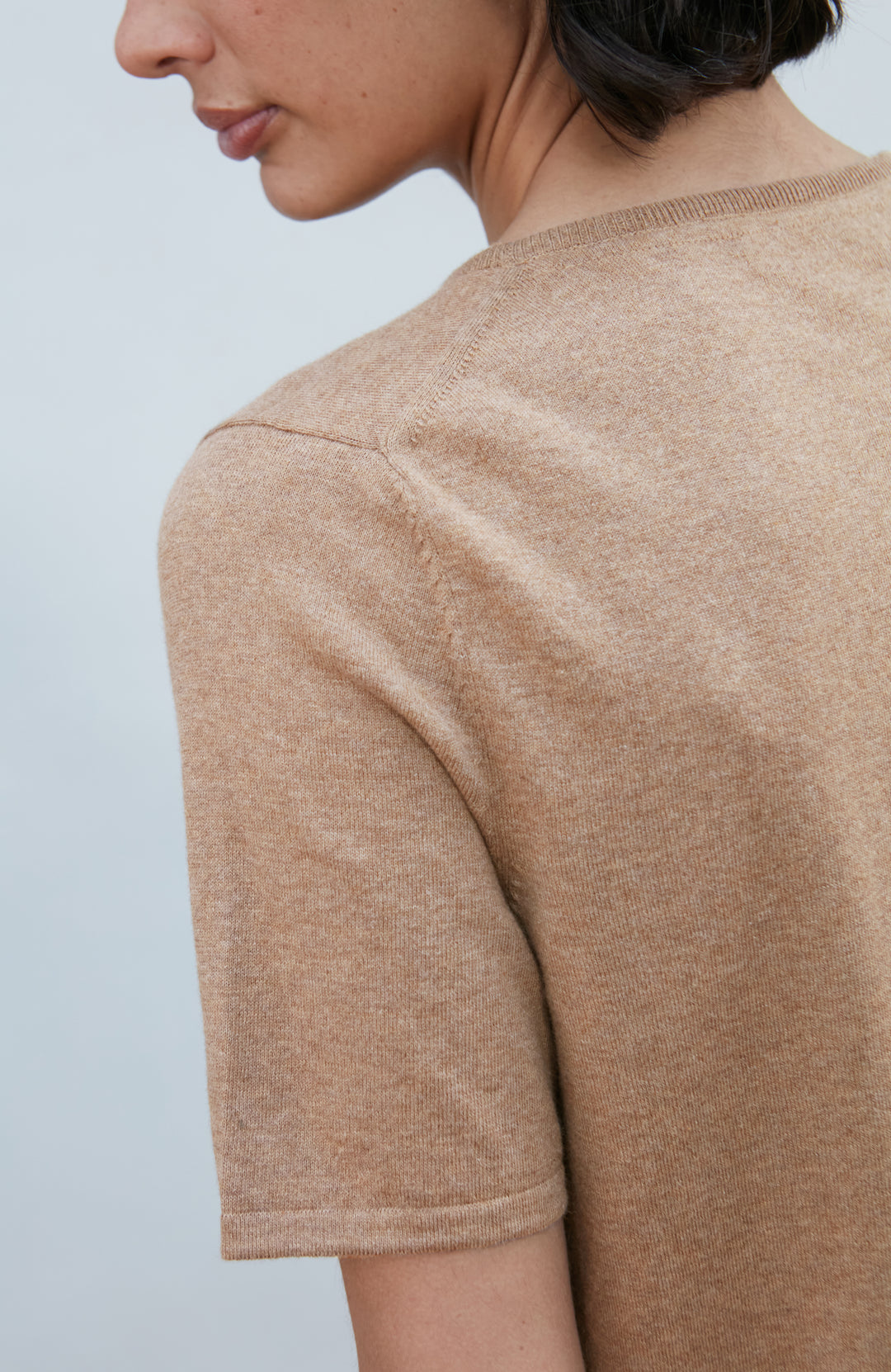 A woman with a short dark bob hairstyle wearing a light brown short-sleeve crewneck knit top by Paul James Knitwear and dark navy pants, standing against a plain light gray background.