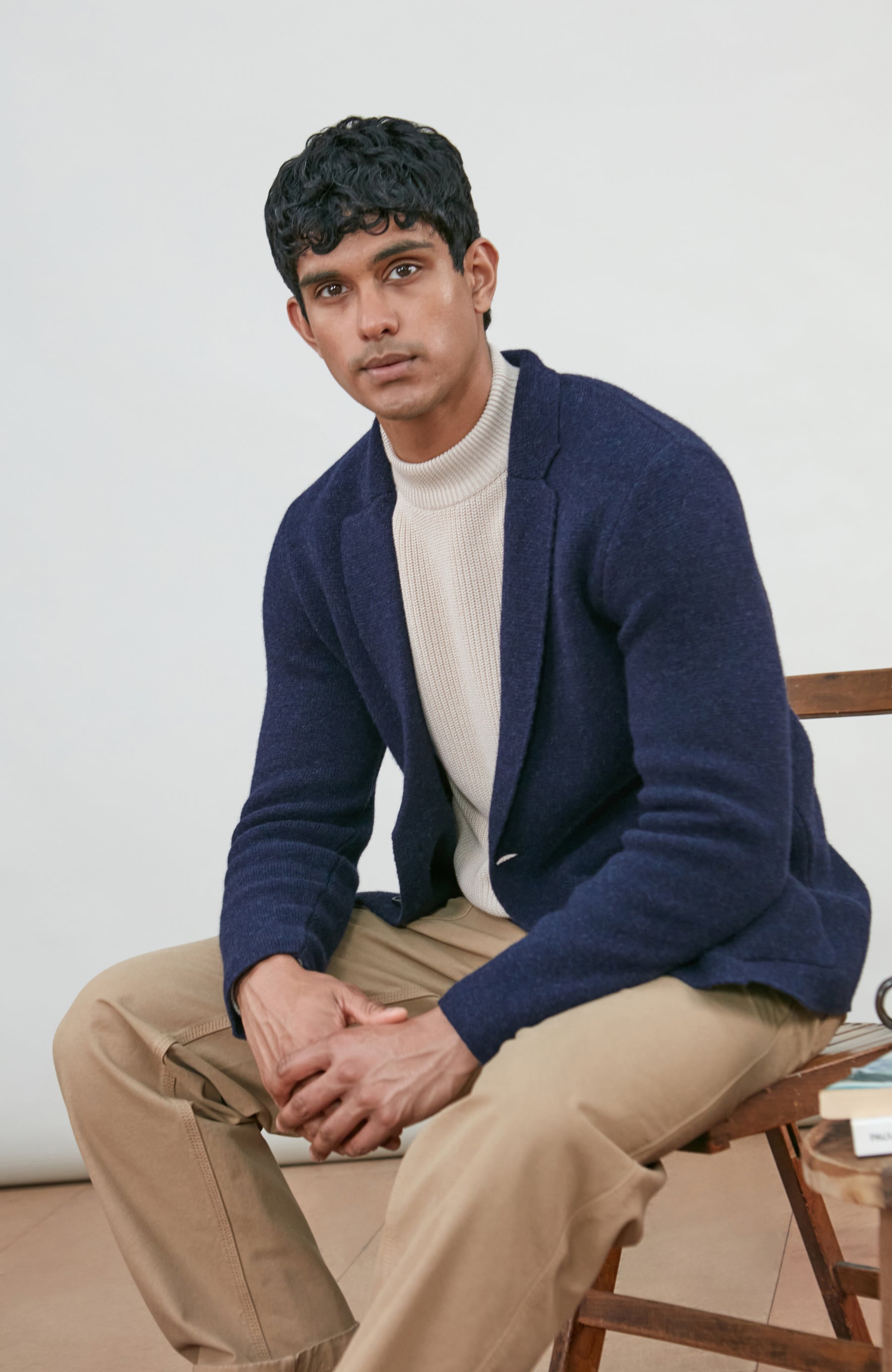 A man sitting on a wooden chair wearing a navy blue wool-knit blazer by Paul James Knitwear over a cream ribbed turtleneck sweater and beige trousers, photographed against a plain light background.