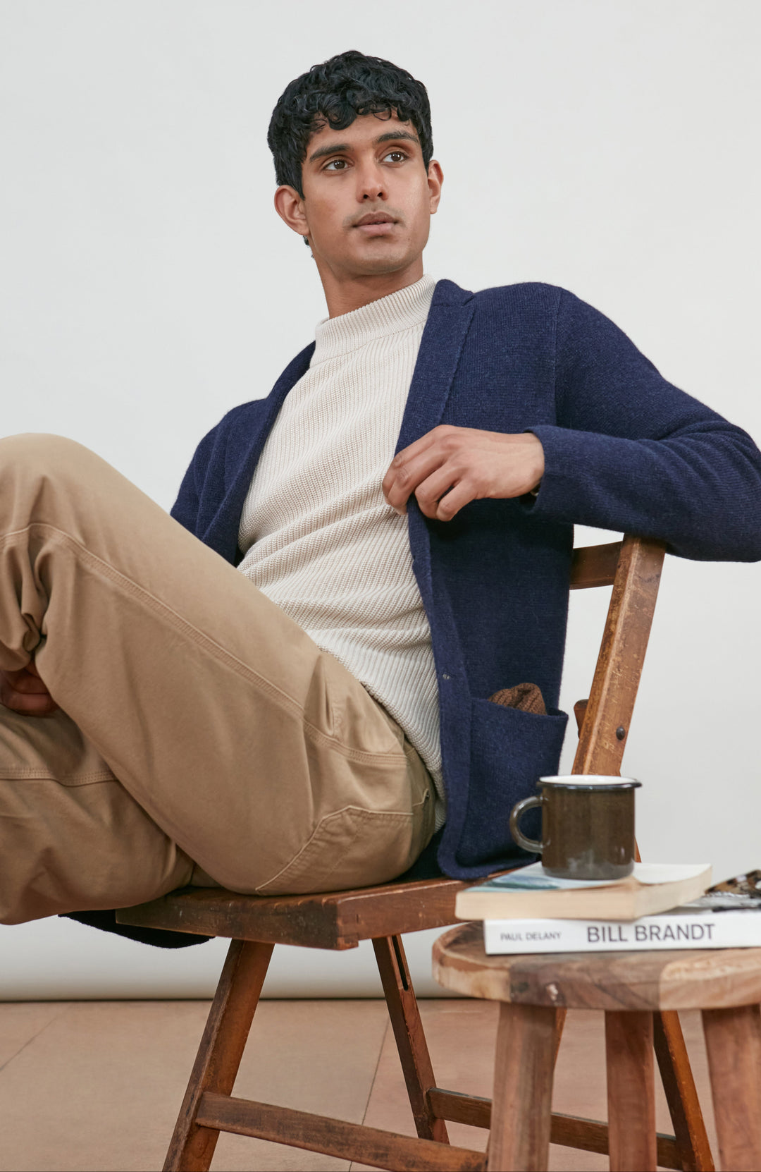 A man sits on a wooden chair, wearing a navy blue wool-knit blazer by Paul James Knitwear over a cream ribbed turtleneck sweater and tan pants. A small wooden side table next to him holds a mug, a book, and a pair of glasses against a plain light background.