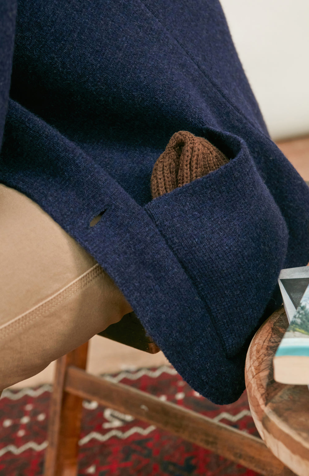 Close-up of a navy blue wool-knit blazer by Paul James Knitwear draped over a person's lap, with a brown knit pocket square peeking out from the pocket, paired with beige trousers.
