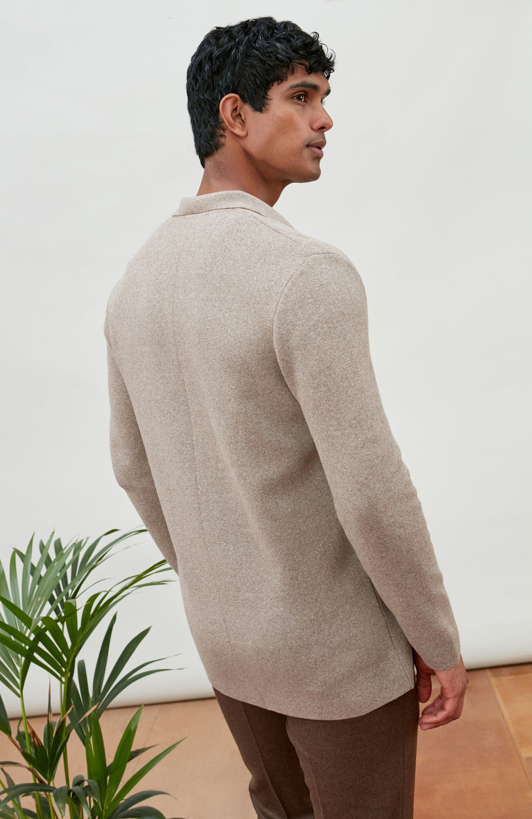 A man wearing a light beige knit sweater and brown trousers, shown from a side-back angle in an indoor setting with a white wall and a green plant.