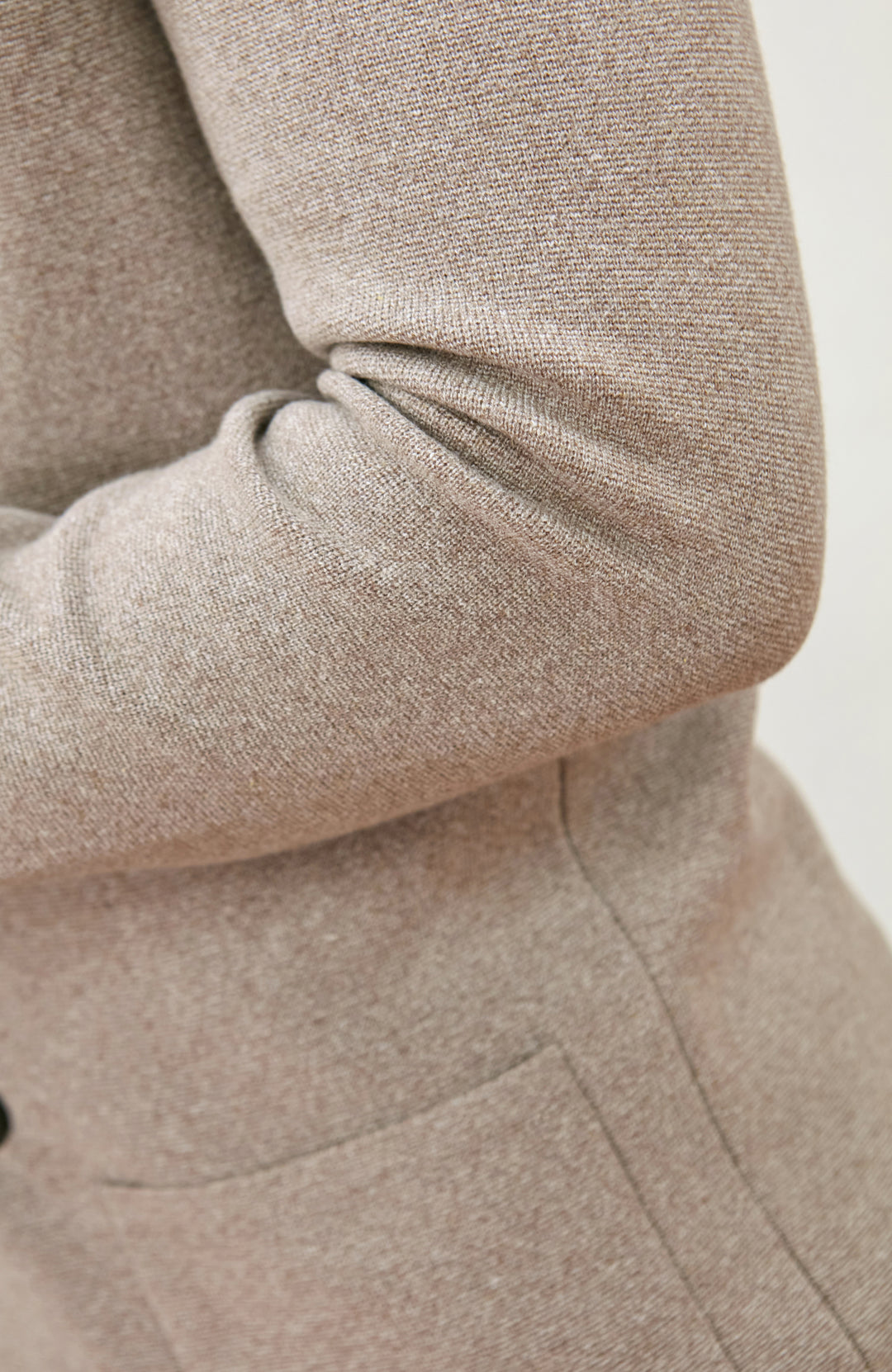 Close-up of a person wearing a textured knit sweater in a warm taupe-beige color, showing the sleeve and upper torso against a plain light background.