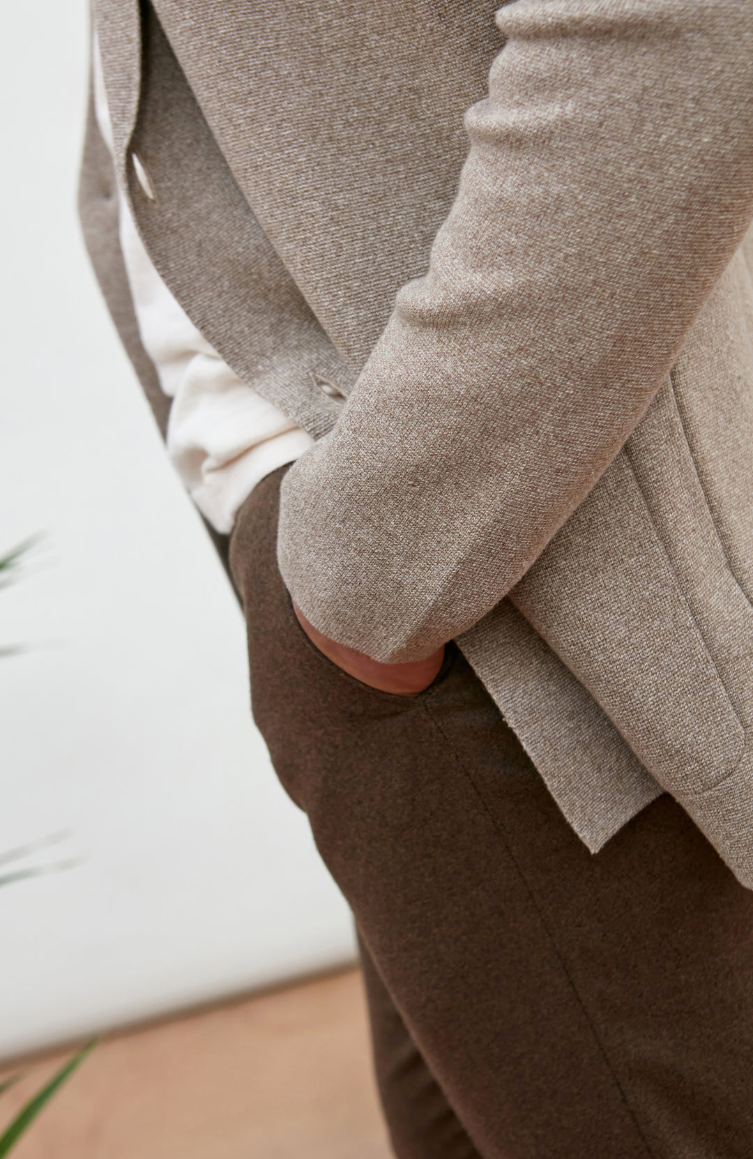 Close-up of a person wearing a light taupe knit blazer over a white shirt and brown trousers, with one hand in a pocket.