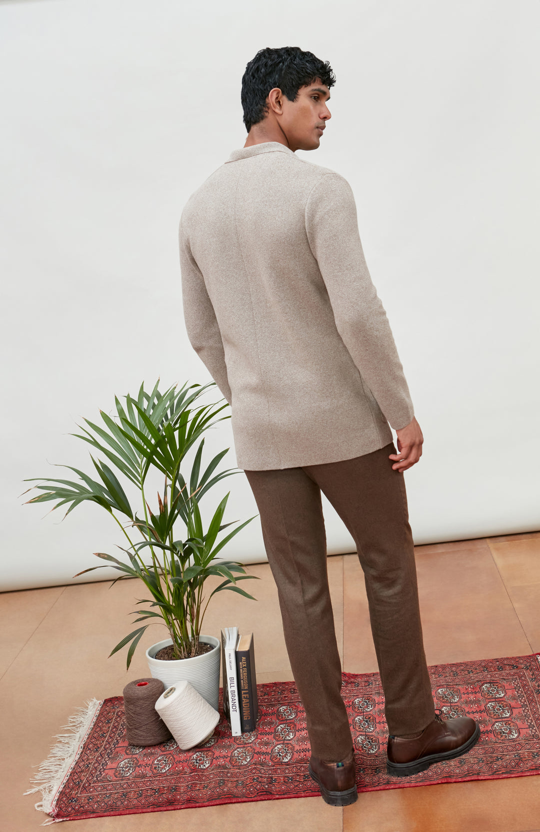 A man stands with his back to the camera wearing a light beige knit sweater and brown trousers, standing on a red patterned rug next to a potted plant.