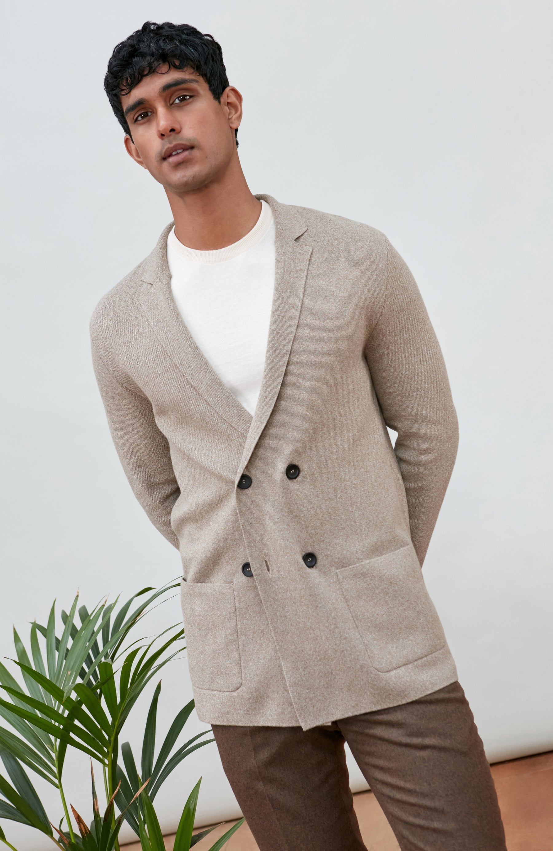 A man wearing a light beige double-breasted knit cardigan by Paul James Knitwear over a white t-shirt and brown trousers, standing in front of a white wall with a green plant in the lower left corner.