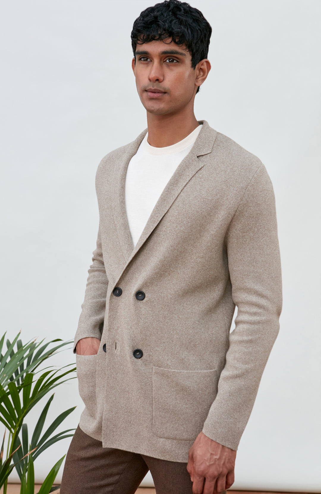 A man wearing a light beige double-breasted knit cardigan by Paul James Knitwear over a white t-shirt and brown trousers