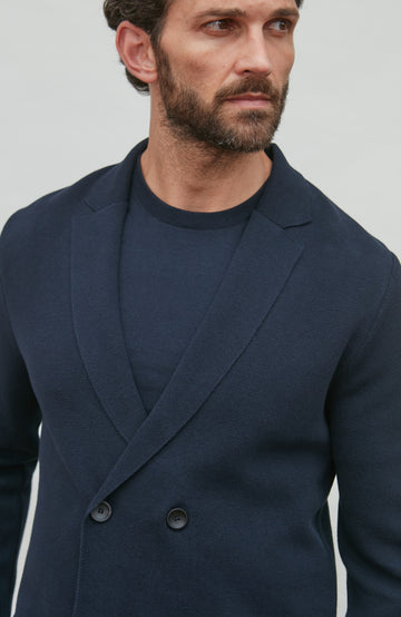 Man wearing a navy blue double-breasted jacket over a black shirt against a light gray background