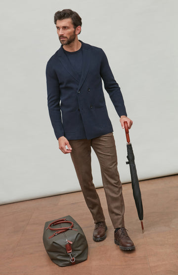 Man wearing navy double breasted blazer holding an umbrella and a bag indoors with a plain background
