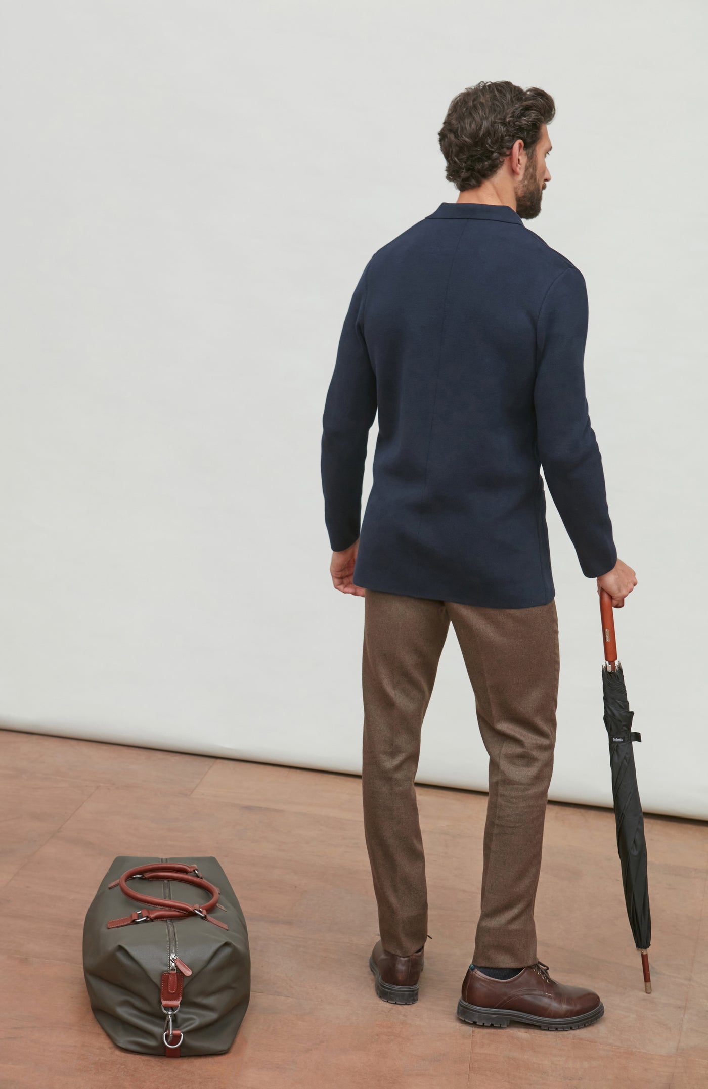 Man in navy double breasted blazer and brown pants holding an umbrella, standing next to a green duffel bag on a light wooden floor.