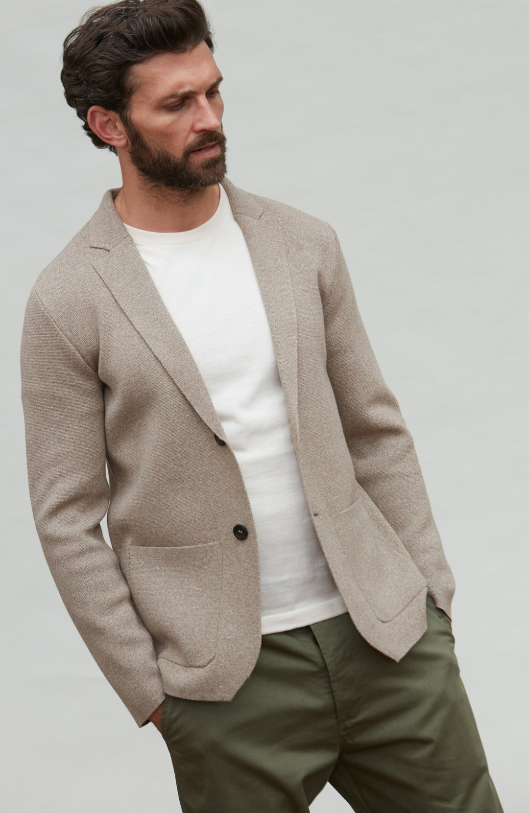 Man wearing a beige knitted blazer over a white shirt and green pants on a plain background