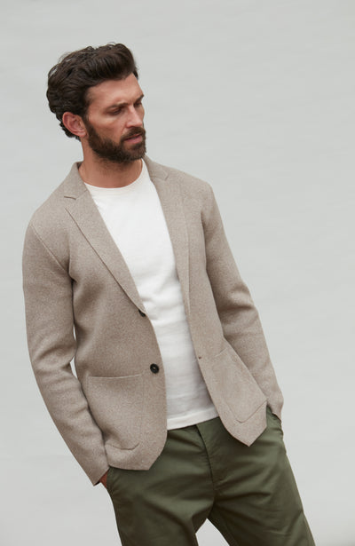 Man wearing a beige knitted blazer over a white shirt and green pants on a plain background