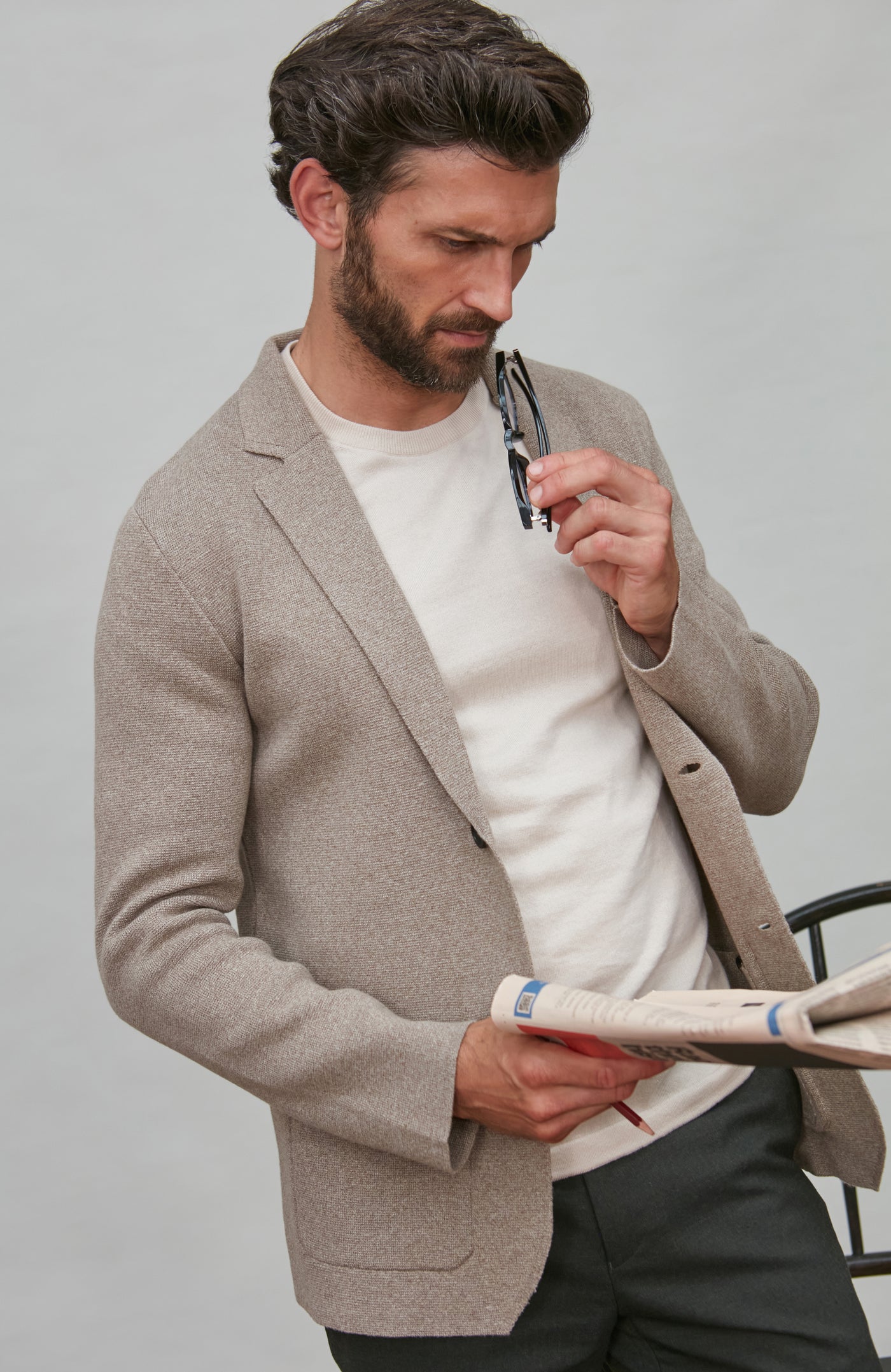 Man wearing a beige knitted blazer over a white t-shirt, holding sunglasses and a newspaper against a plain background