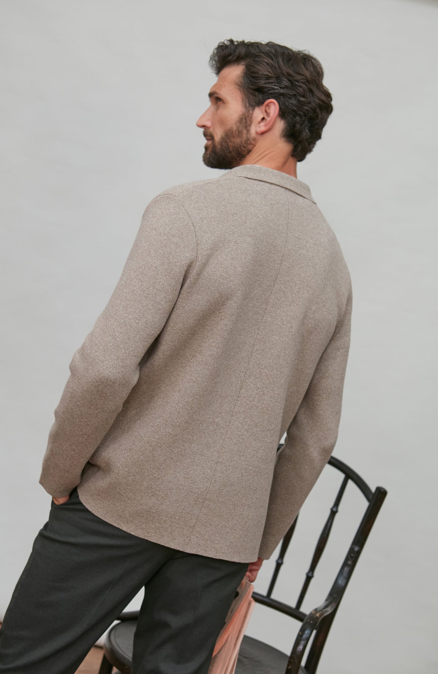 Man wearing a beige paul james knitted blazer and dark pants sitting on a chair against a plain background