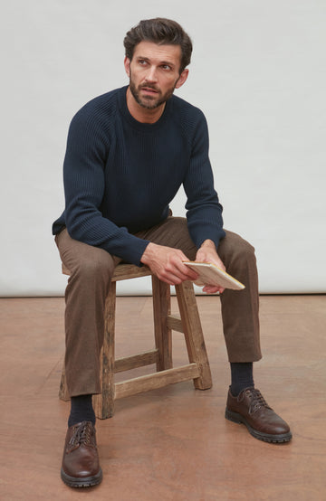 Man sitting on a wooden stool wearing a navy fisherman sweater, brown pants, and brown shoes by Paul James