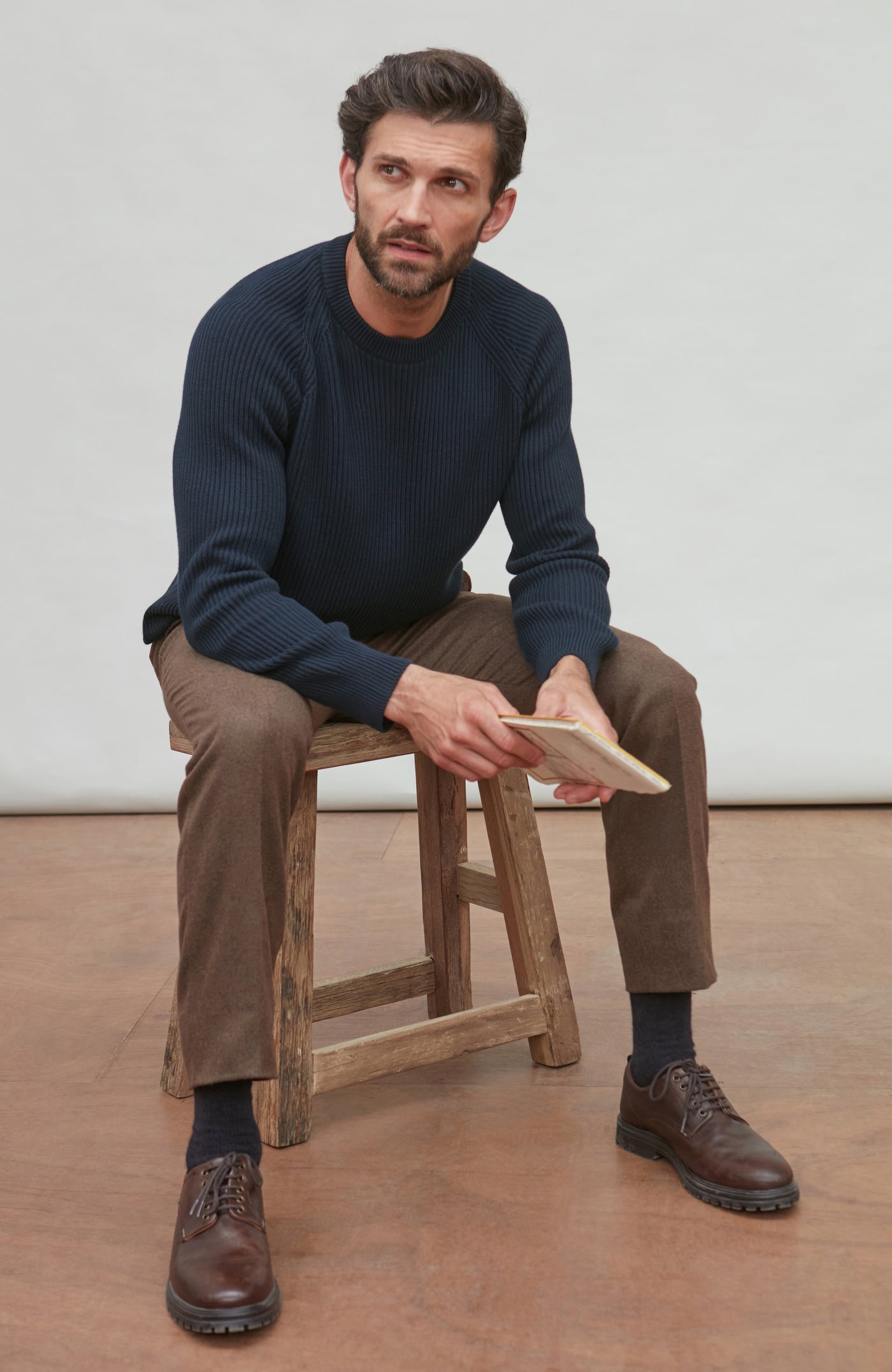 Man sitting on a wooden stool wearing a navy fisherman sweater, brown pants, and brown shoes.