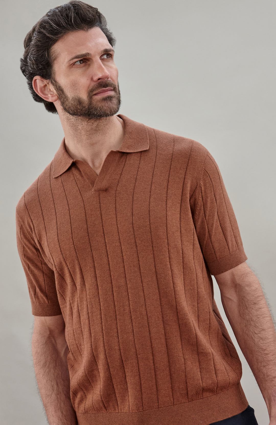 A man wearing a short-sleeve knit polo shirt in a warm rust brown color, shown from the chest up against a plain light gray studio background.