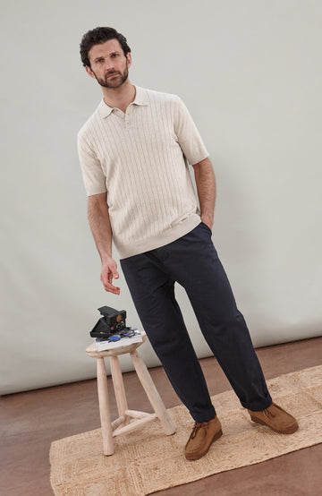 A man wearing a cream short-sleeve rib-knit cotton polo shirt by Paul James Knitwear, navy trousers, and brown suede shoes, standing in a studio setting next to a small wooden stool.
