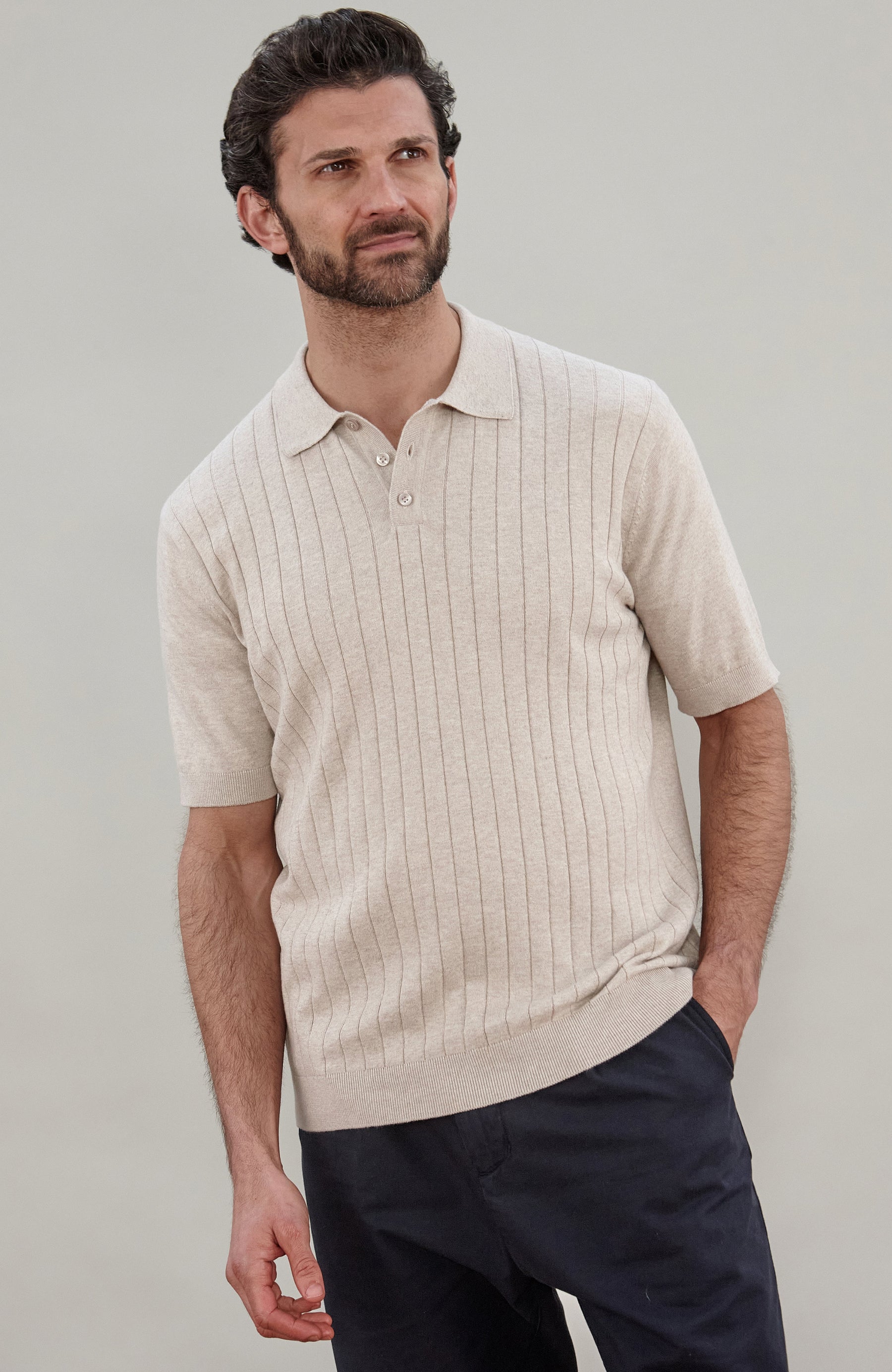 A man wearing a short-sleeve rib knit cotton polo shirt by paul james knitwear in a light beige color with vertical ribbed texture, paired with dark navy pants, against a solid light gray background.