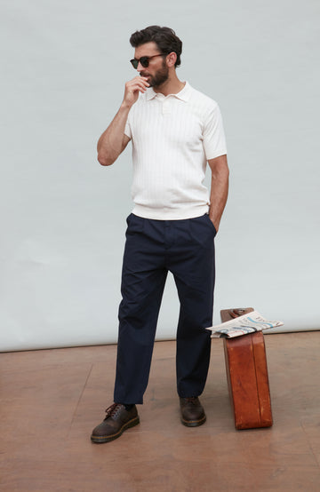 Man in white polo shirt and navy pants standing next to a suitcase against a plain background