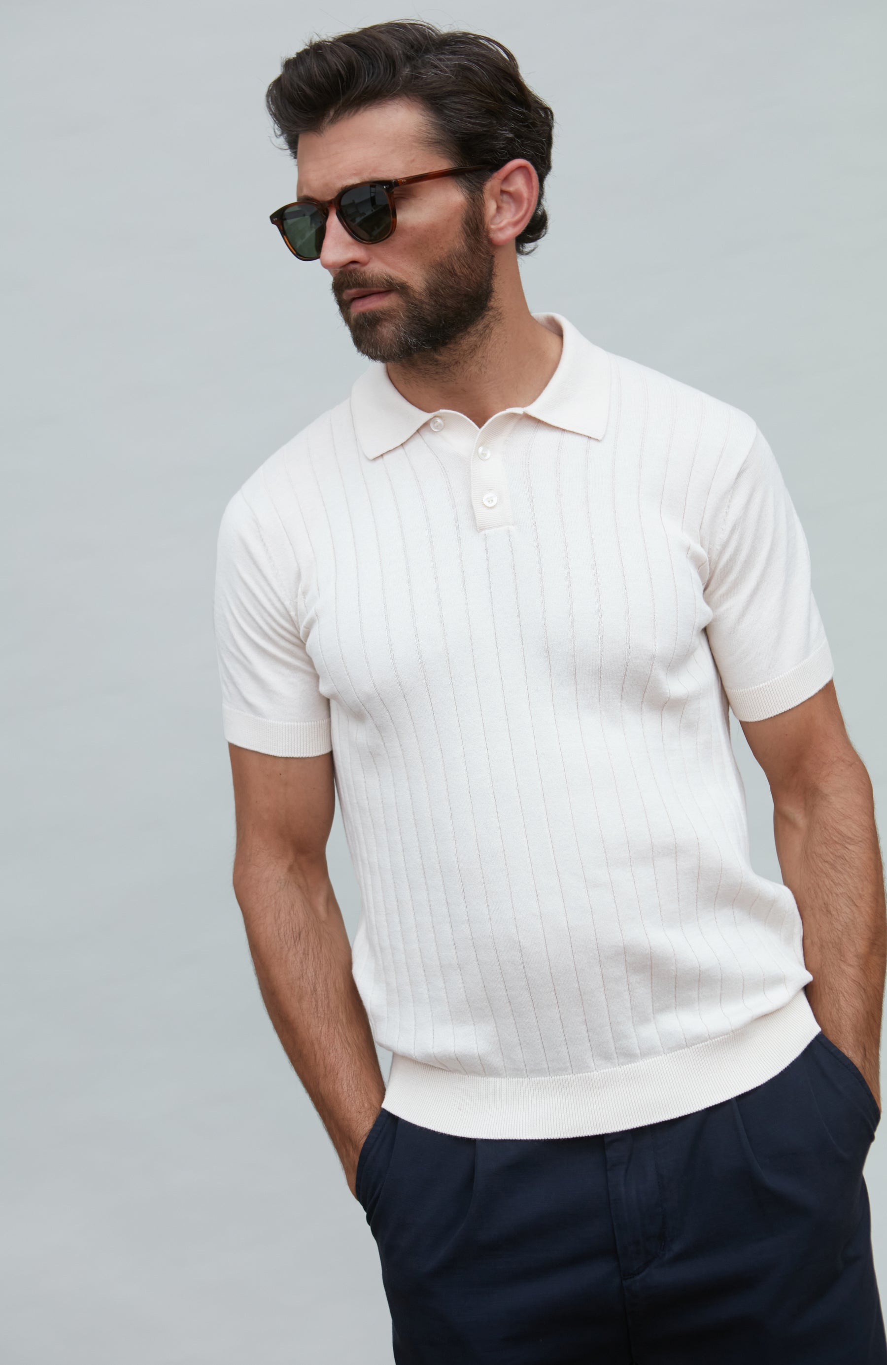 Man wearing a white ribbed polo shirt and dark pants against a gray background