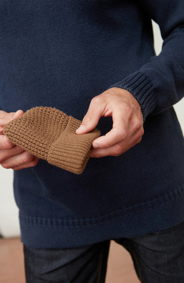 Person holding a brown knitted beanie against a dark blue sweater background by Paul James