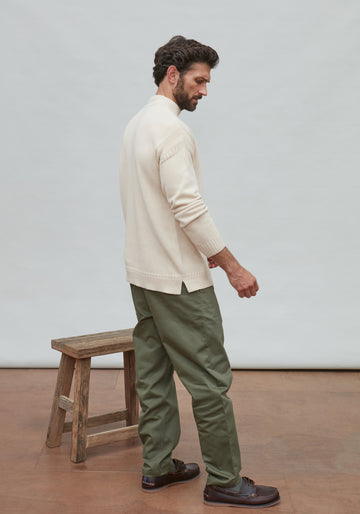 Man wearing a cream guernsey sweater and green pants standing next to a wooden stool against a plain background