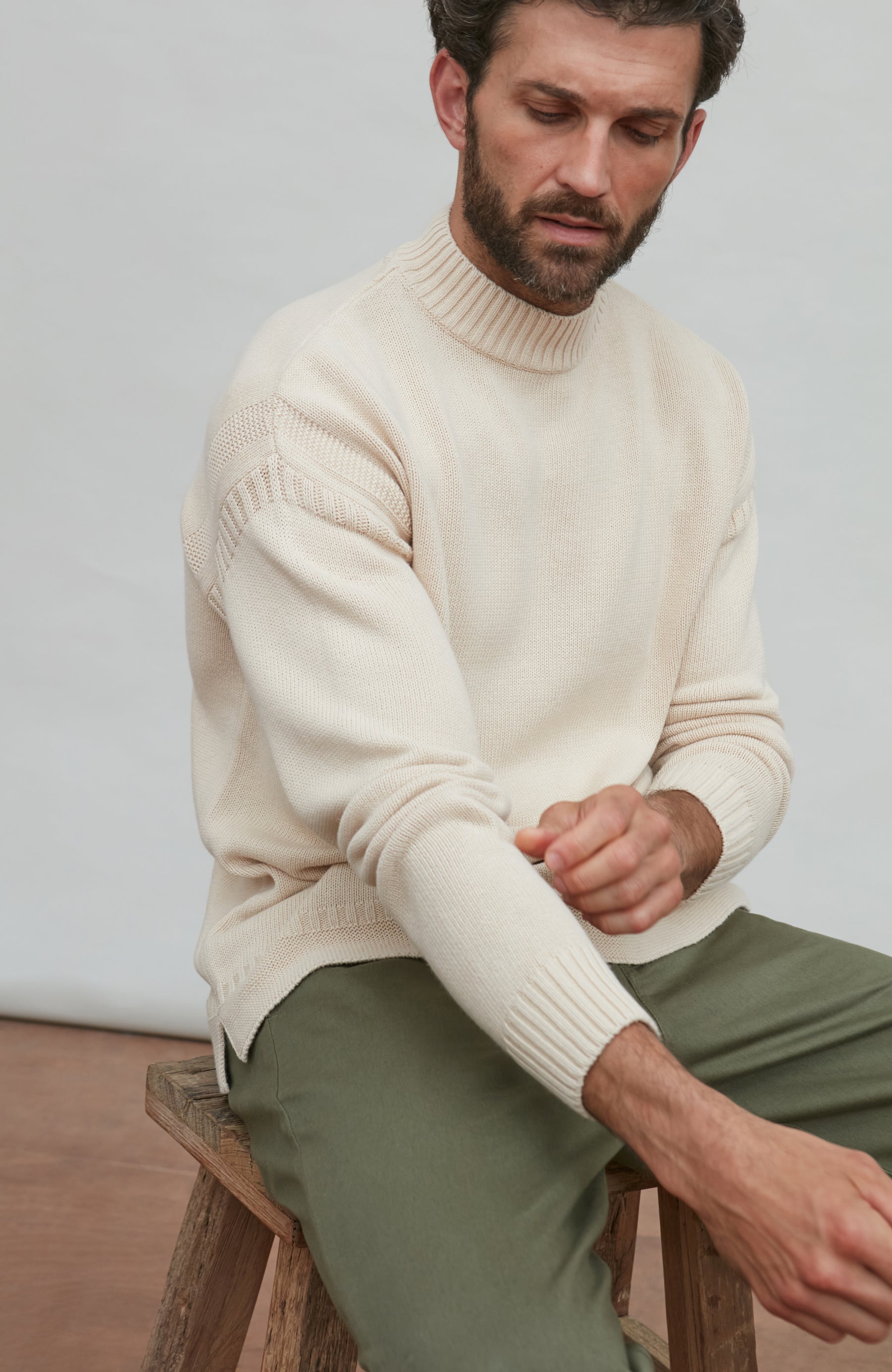 Man wearing a cream guernsey sweater and green pants sitting on a wooden stool.