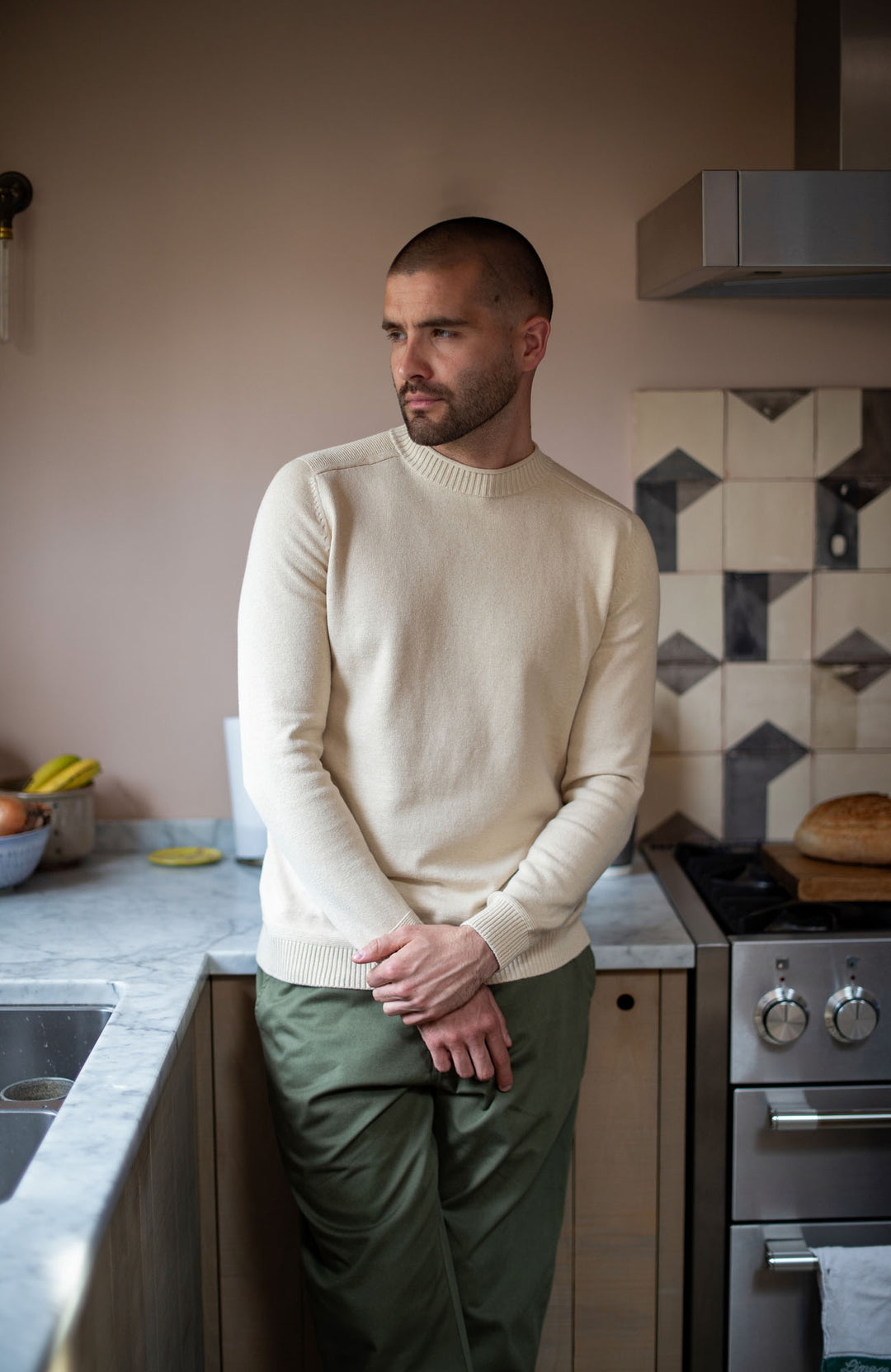 Man standing in a kitchen wearing a beige sweater and green pants by Paul James