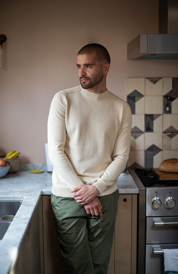 Man standing in a kitchen wearing a beige sweater and green pants by Paul James