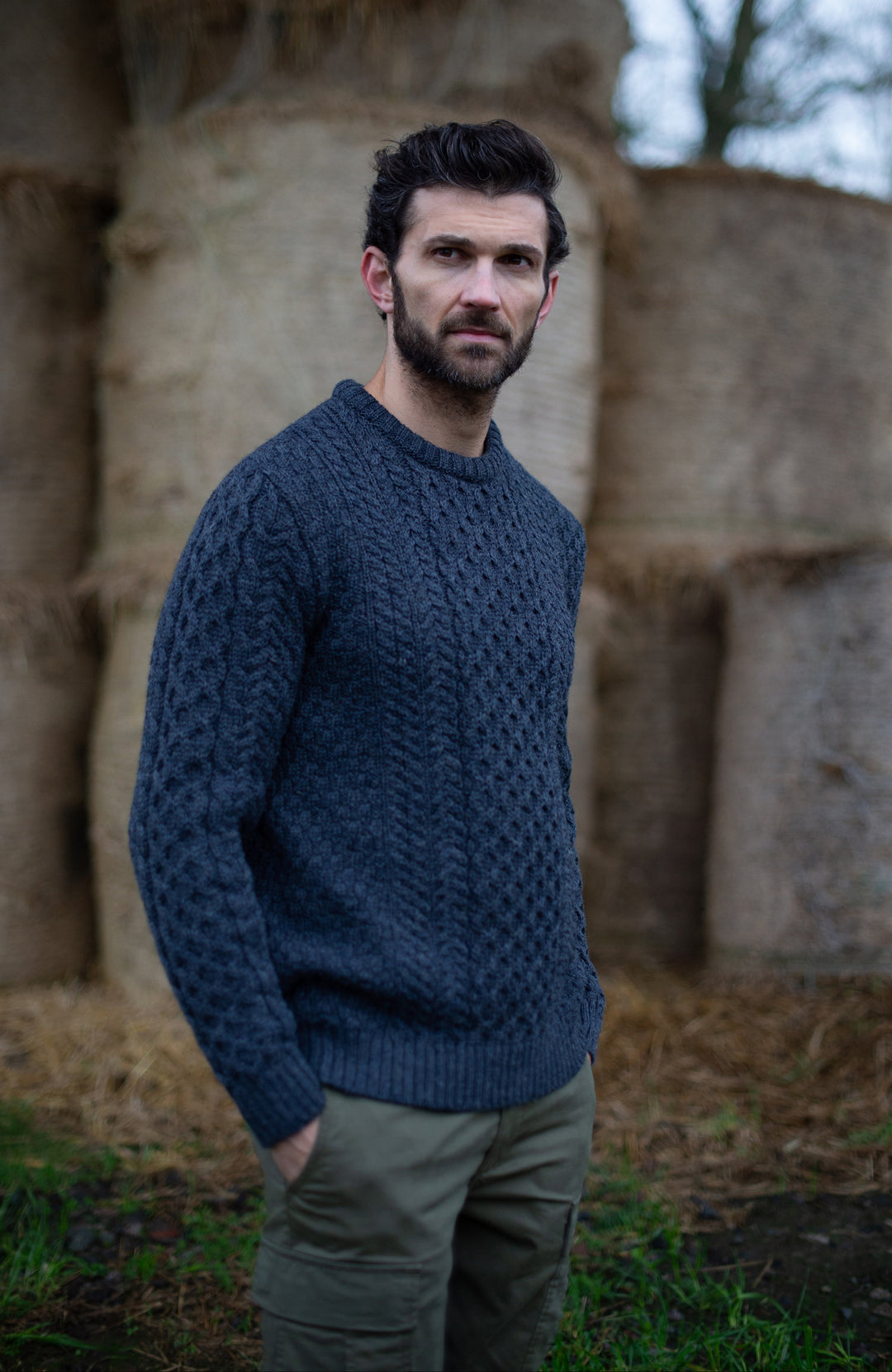 Man wearing a blue knitted British wool cable sweater ny Paul James Knitwear standing in front of stone structures.