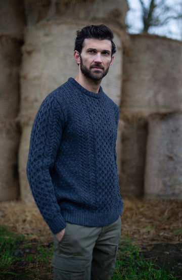 Man wearing a blue knitted British wool cable sweater ny Paul James Knitwear standing in front of stone structures.