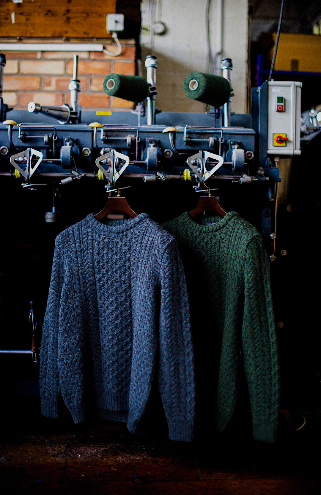 Two cable-knit crewneck British wool Paul James sweaters hanging on a machine in a workshop. The sweater on the left is a muted slate blue, and the sweater on the right is a forest green.