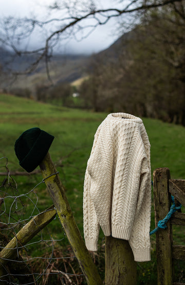 White knitted british wool sweater and black hat hanging on a wooden fence with a scenic background By Paul James
