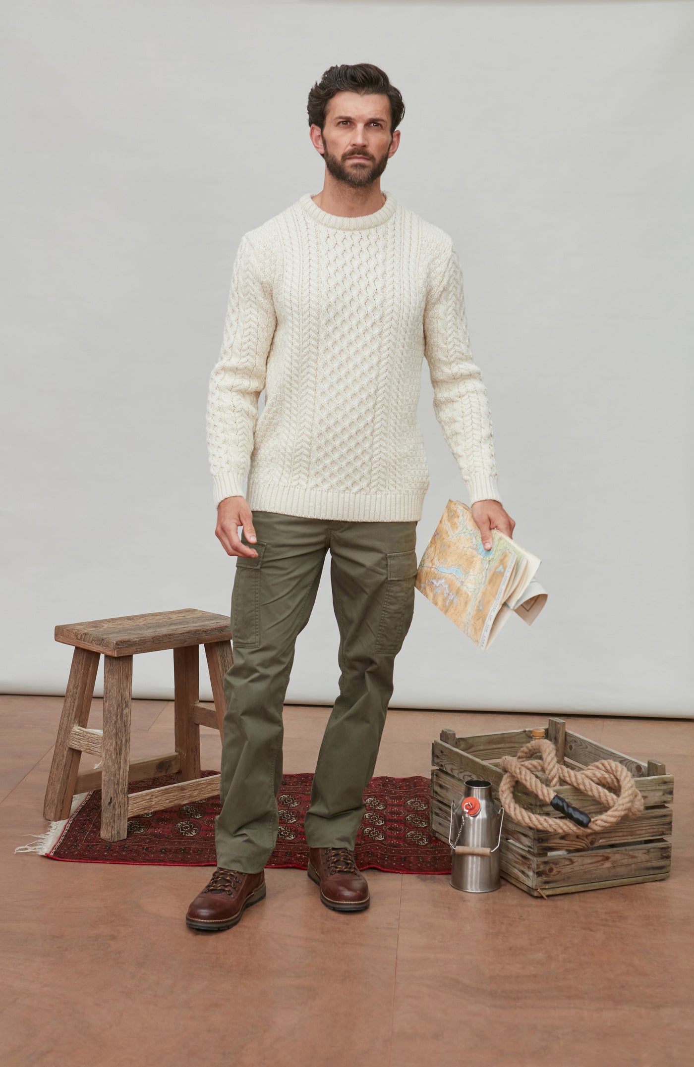 Man wearing a cream cable british wool sweater and green pants standing in a room with a wooden stool, crate, and rug.