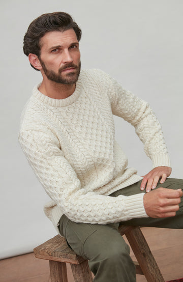 Man wearing a white textured cable British wool sweater sitting on a wooden stool against a plain background By Paul James