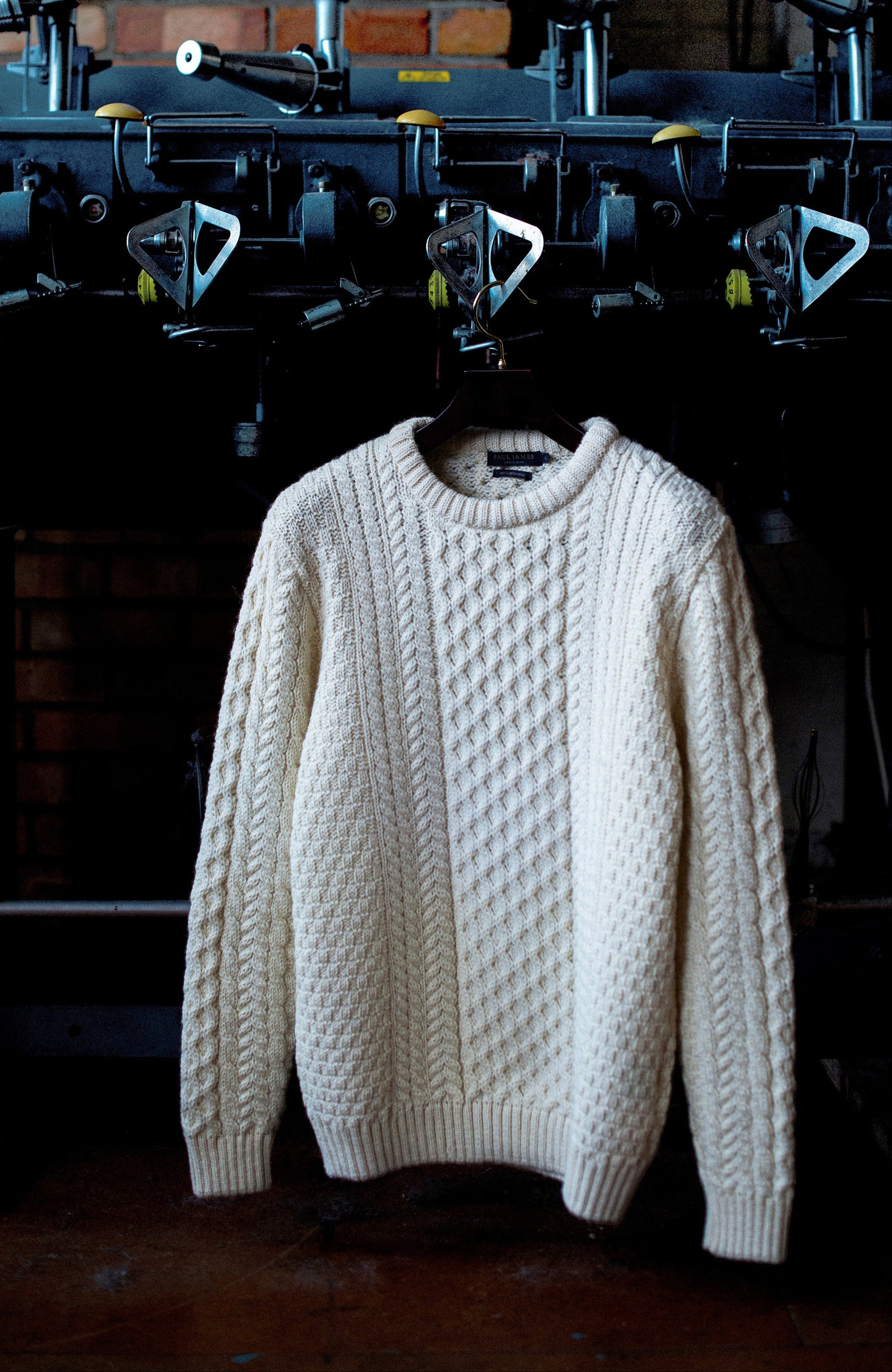 An off-white British wool cable-knit crewneck sweater by Paul James Knitwear hanging on a hanger in front of a dark, vintage industrial press machine.