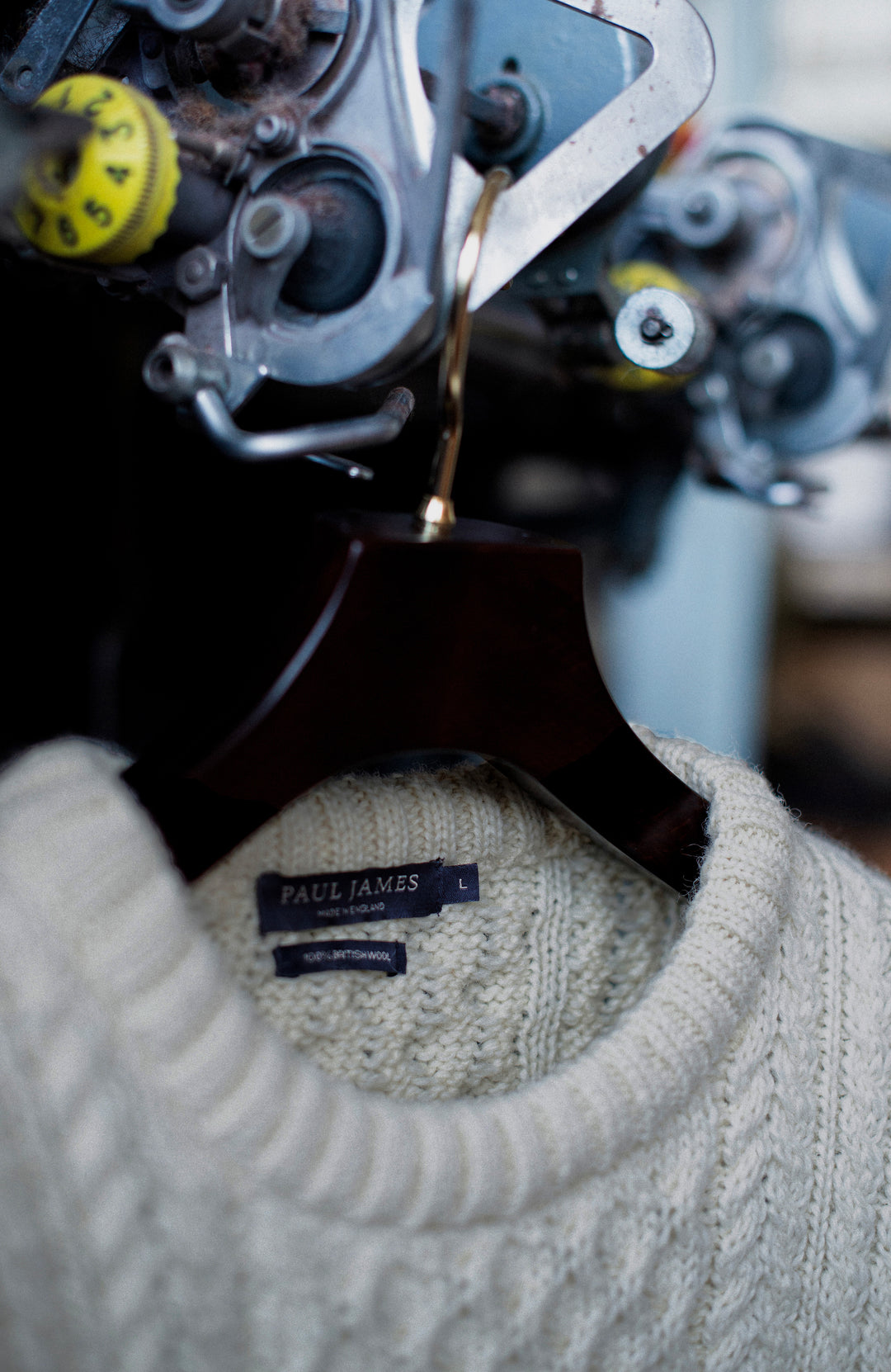 A close-up of a cream-colored British wool cable-knit Paul James sweater hanging on a dark wooden hanger, with a visible Paul James neck label. The background is softly blurred, showing vintage industrial machinery.