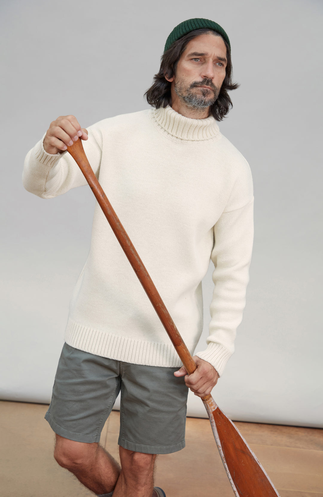 A man wearing a cream wool submariner ribbed turtleneck sweater, gray shorts, and a dark green beanie, holding a wooden paddle against a light gray studio background. Made in England by Paul James Knitwear.